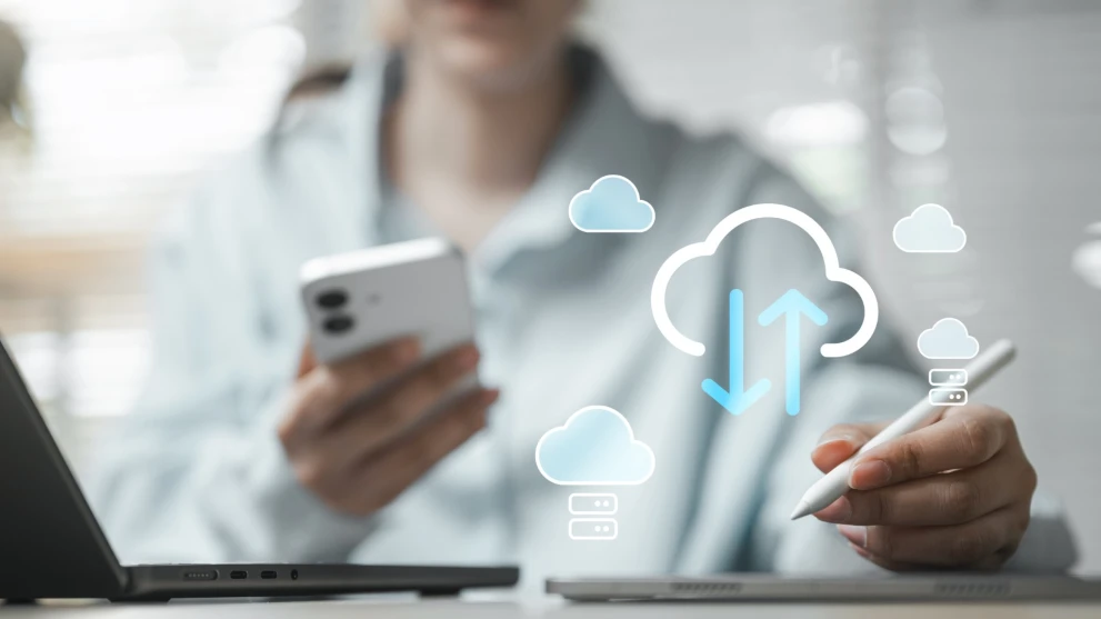 Das Bild zeigt eine moderne Nutzung von Cloud-Technologie mit Smartphone, Tablet und Laptop im vernetzten Arbeitsalltag. Visualisierte Cloud-Symbole mit Upload- und Download-Pfeilen stehen für Datensynchronisation, Cloud-Speicher und den sicheren Zugriff auf digitale Inhalte über mehrere Geräte.