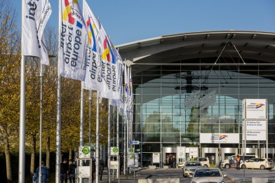 Entrance area of Messe München with flags of the 'inter airport Europe' trade fair and taxis waiting in front of the modern glass building on a sunny day.