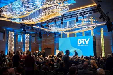 View into the event hall of the DRV Capital Congress 2025 in Berlin: Numerous participants wait in front of the large stage with the DRV logo for the program to begin. The ISO-Gruppe is on site as a partner and contributes its expertise in the field of tourism IT and digitalization.