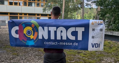 Person hält ein Banner der Firmenkontaktmesse CONTACT 2025 mit Logo und URL contact-messe.de auf dem Campus der FAU Erlangen-Nürnberg.